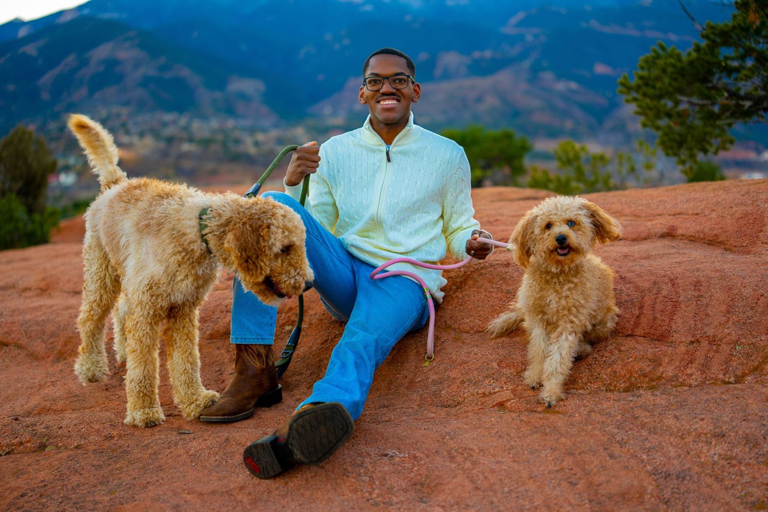 A goldendoodle dog and person sit together on red rocks against a backdrop of mountains and blue sky.