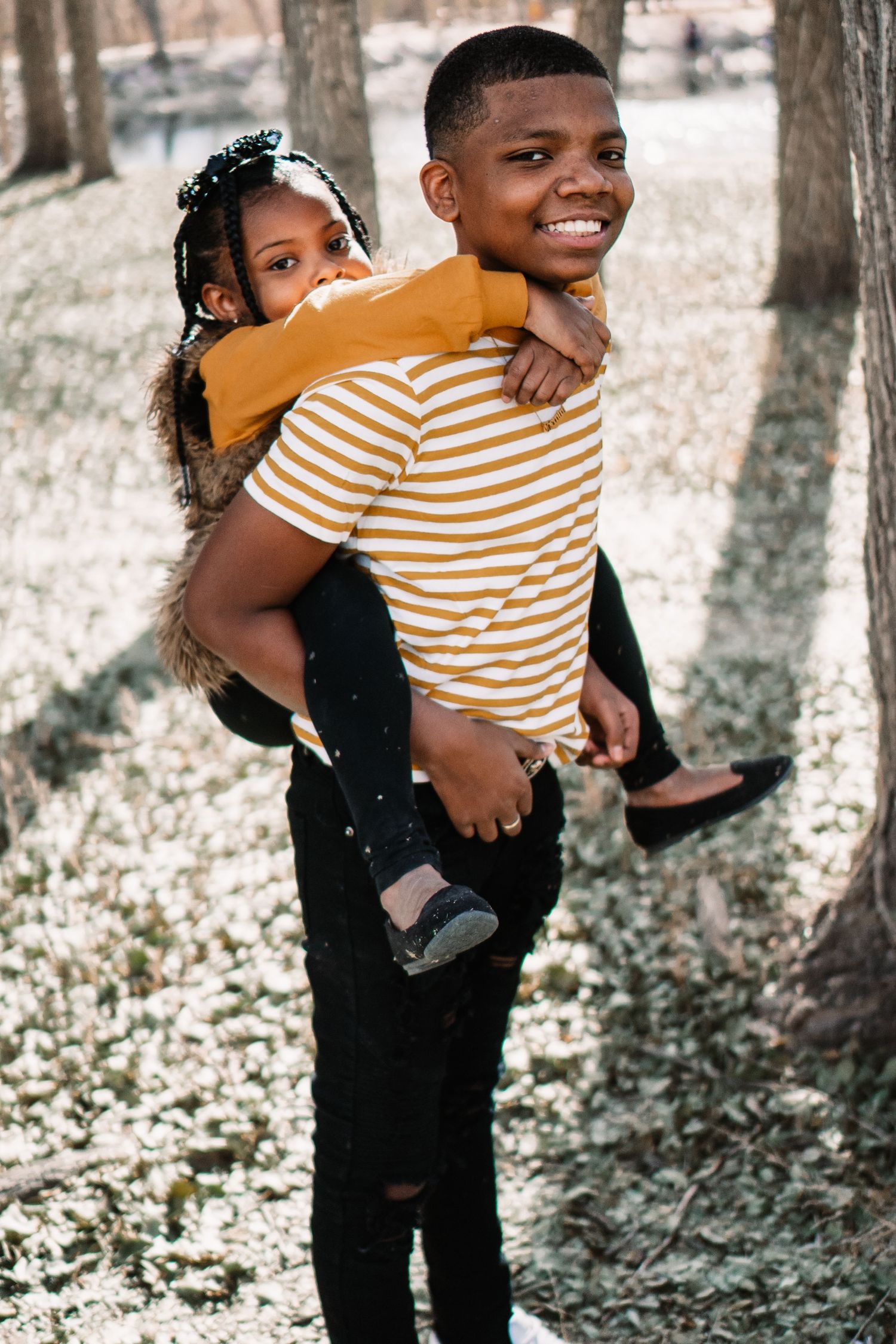 Two young friends sharing a playful piggyback ride outdoors in a wooded setting.