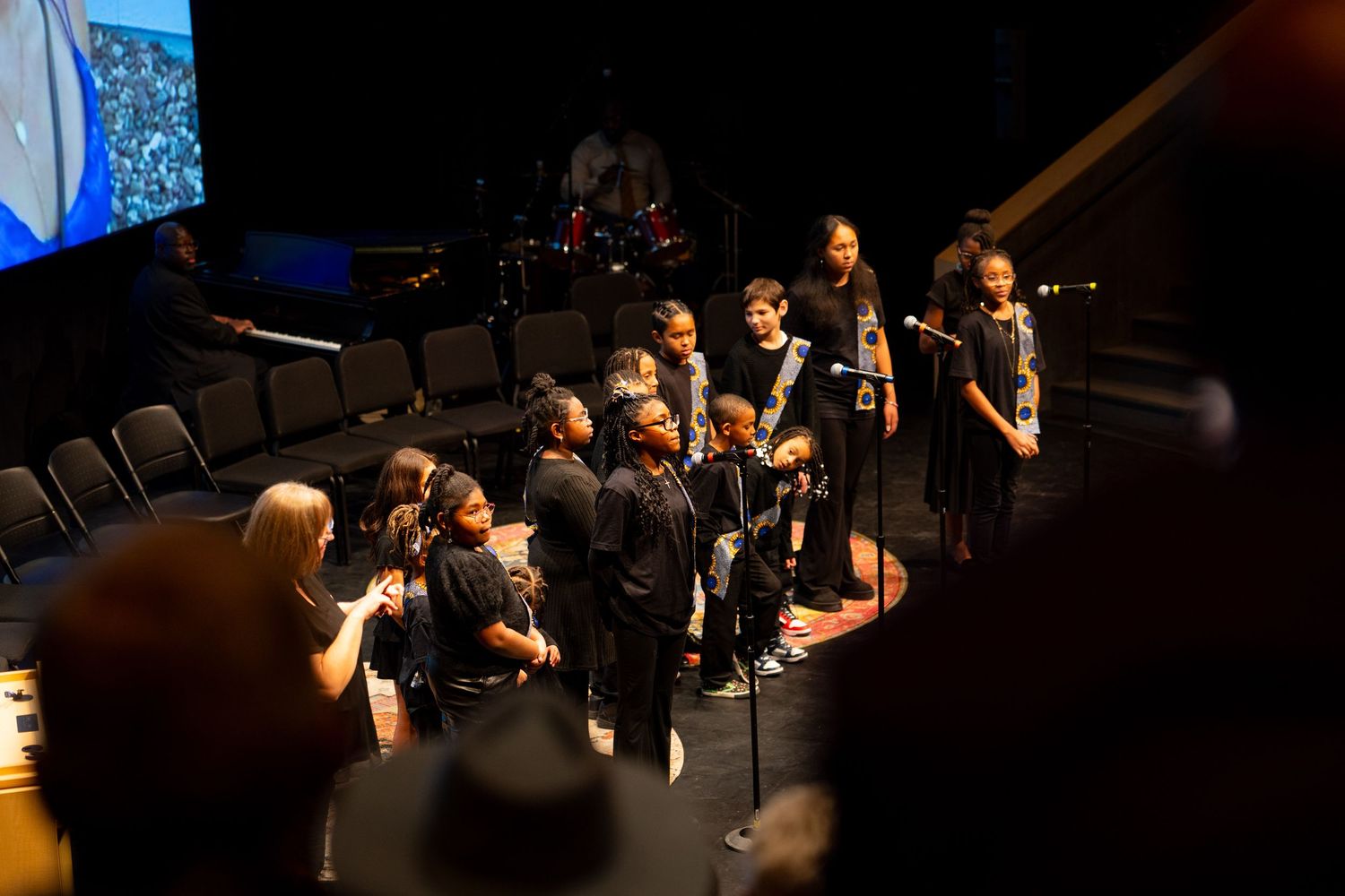Children choir group performing on stage in a darkened auditorium during a formal event.