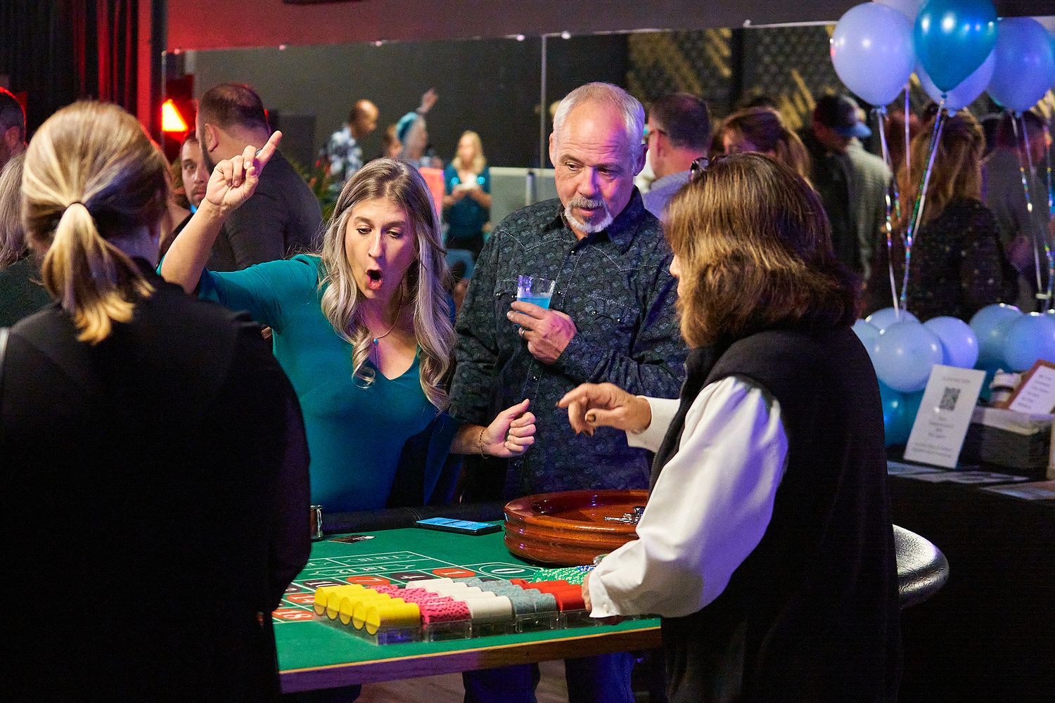 Casino players gather around a table game with colorful balloons in the background at an evening event.