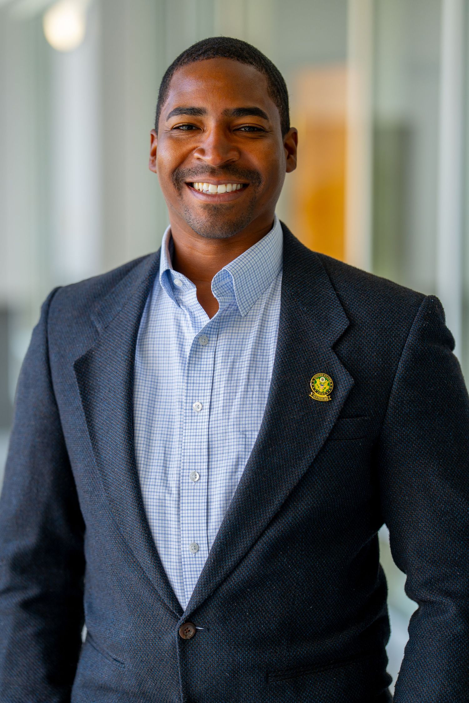 Professional headshot of someone wearing a light blue button-down shirt and dark blazer with a pin on the lapel.