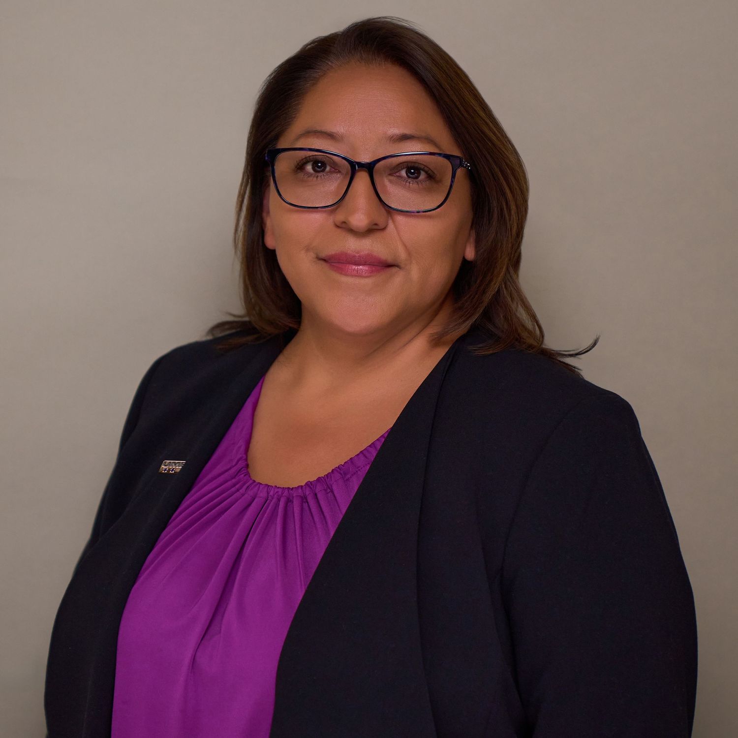 Professional headshot of someone wearing glasses and a purple blouse with a dark blazer against a neutral background.