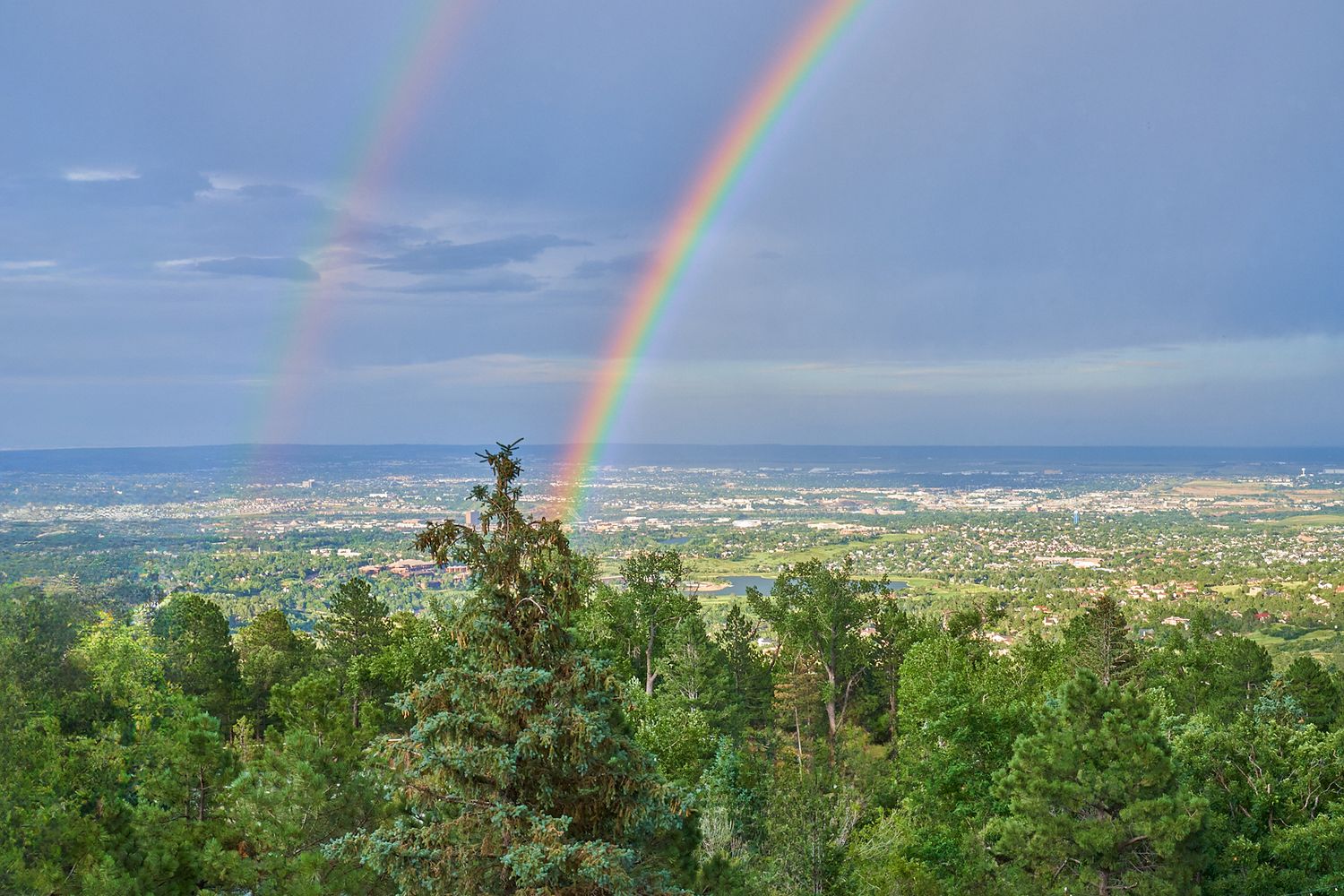 Double rainbow arches over Colorado Springs cityscape viewed from a mountain overlook.