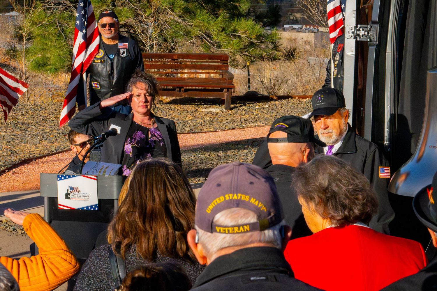 A group gathers outdoors near American flags and benches in a scenic desert setting during a sunny day.