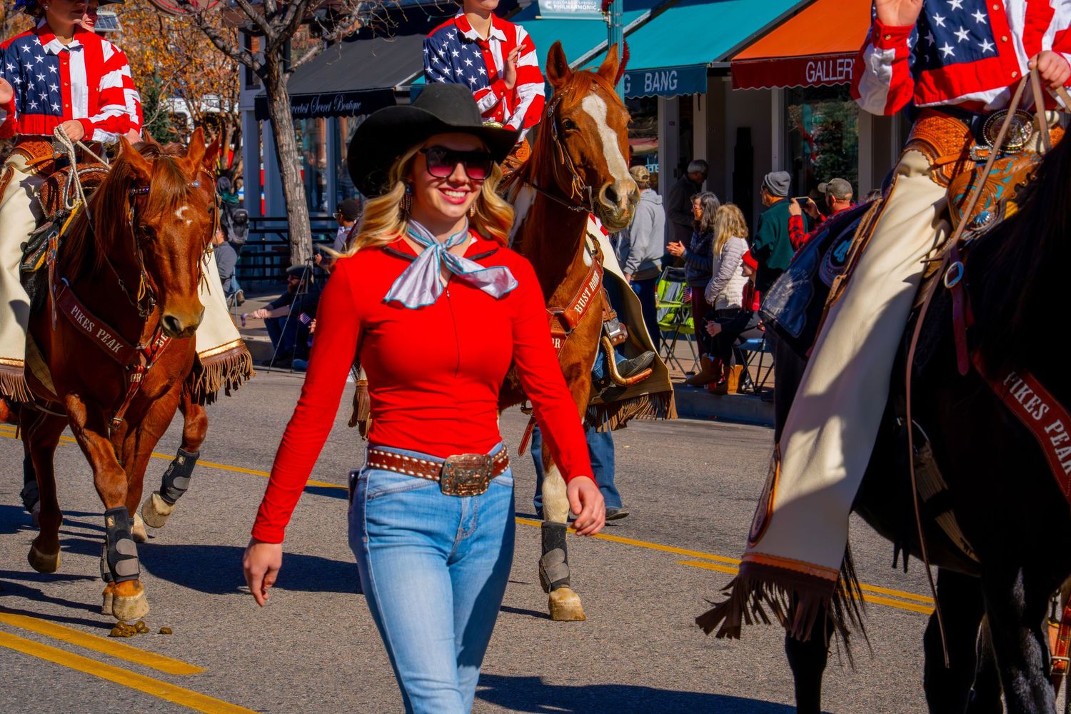 Parade participant in red western shirt and jeans walks alongside horses with American flags in background.