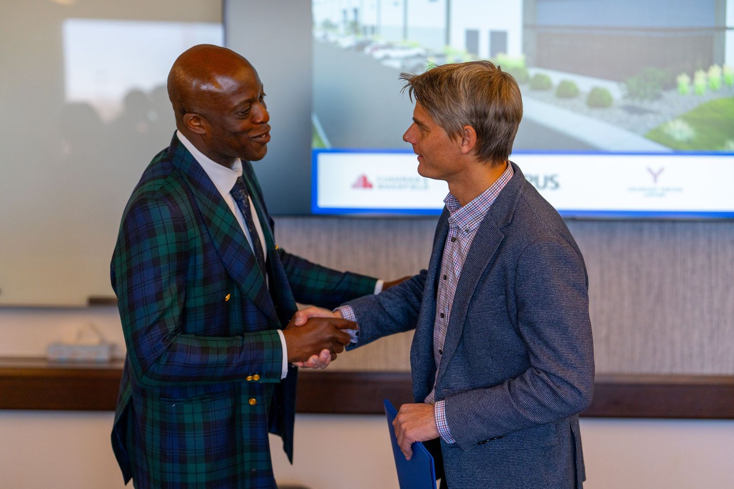 Two businessmen in suits shake hands in front of a projected building rendering at a corporate event.