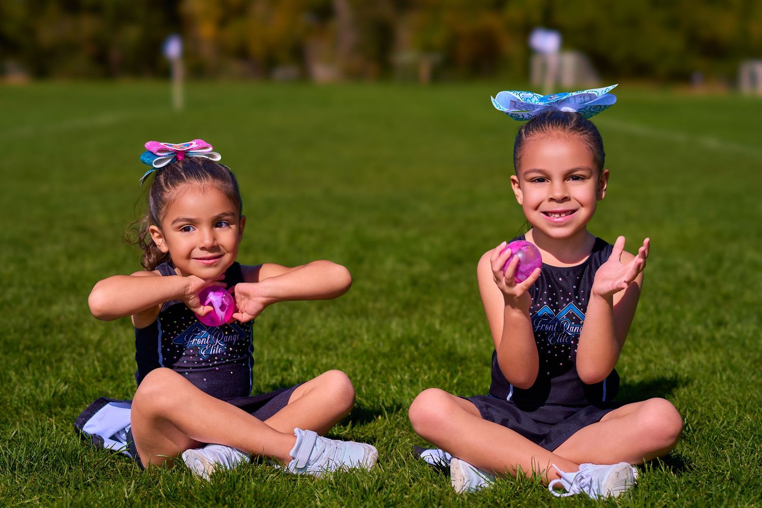 Two young dancers in matching blue outfits pose playfully on grass with bows in their hair.