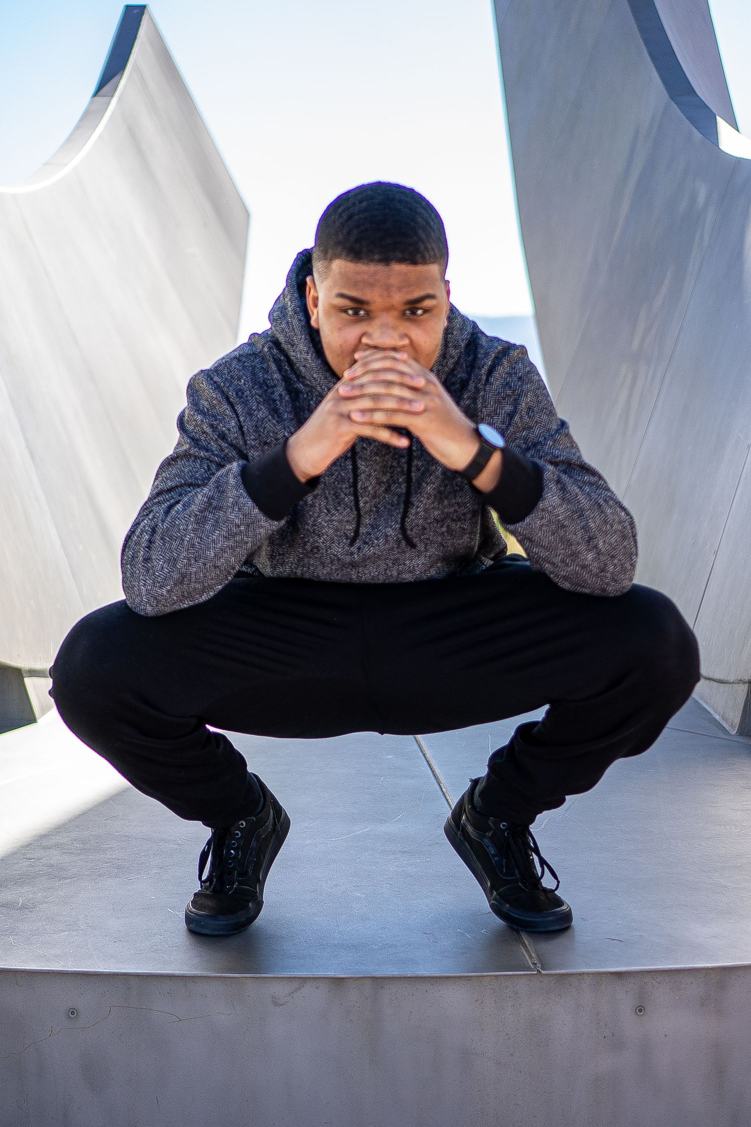 Person in grey hoodie crouching against white architectural background in urban setting.
