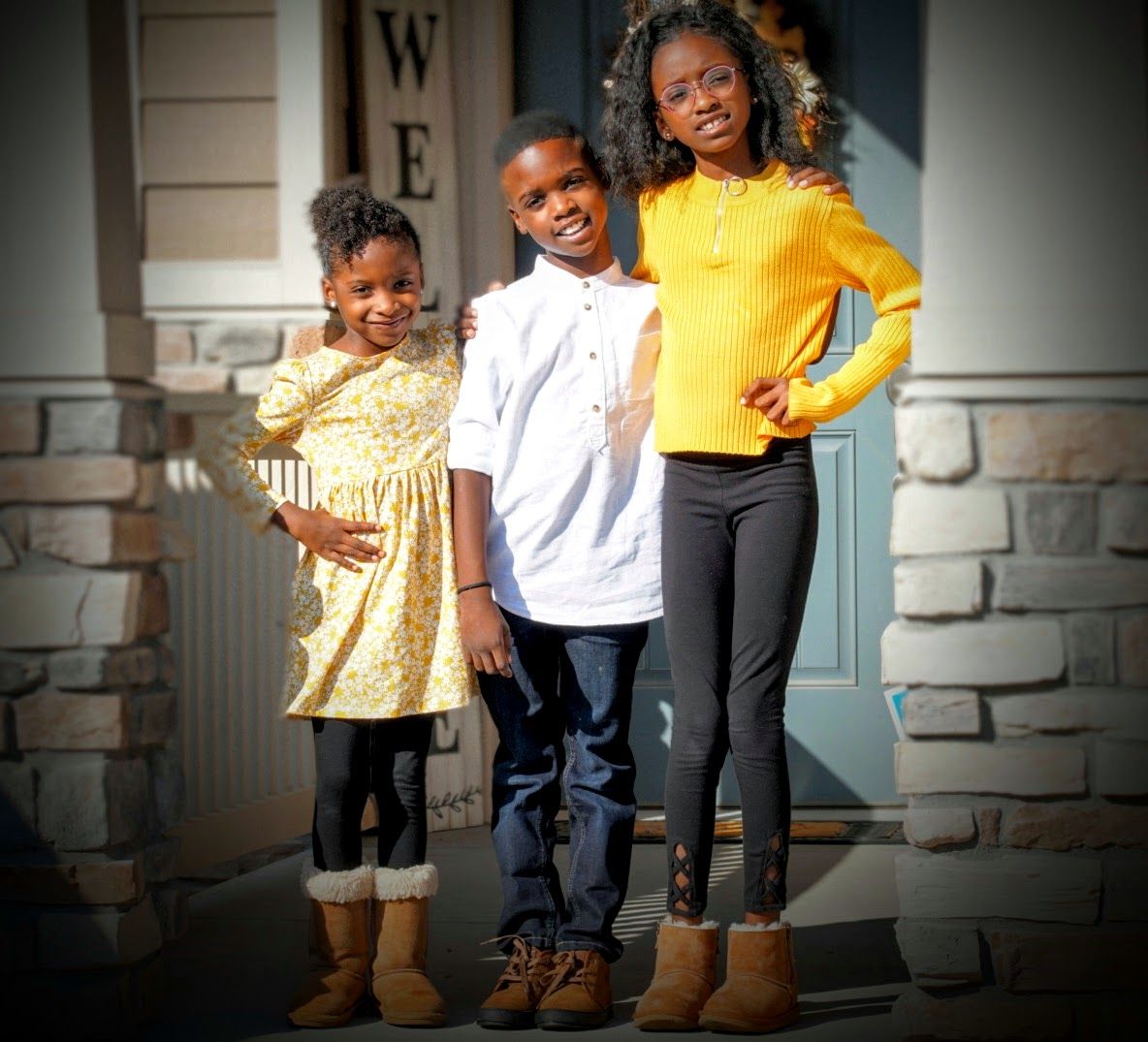 Three individuals pose together on a front porch with stone accents and a welcome sign.