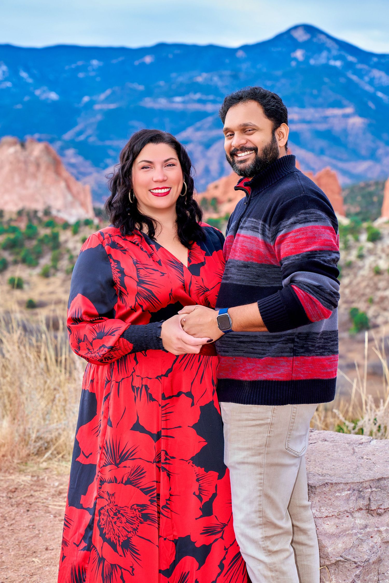 Couple poses together on trail with woman in red floral dress against mountain backdrop.