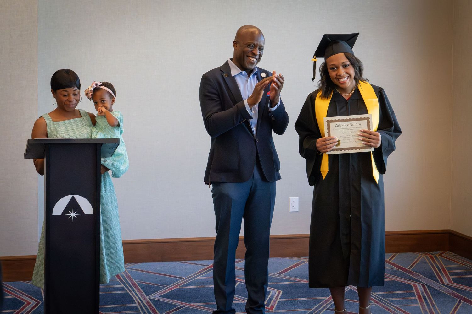 Graduate in cap and gown holds diploma certificate while man applauds at indoor graduation ceremony.
