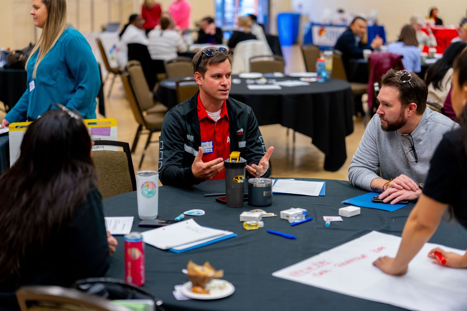 Professionals engage in discussion at a conference workshop, seated around black tables with papers and drinks.
