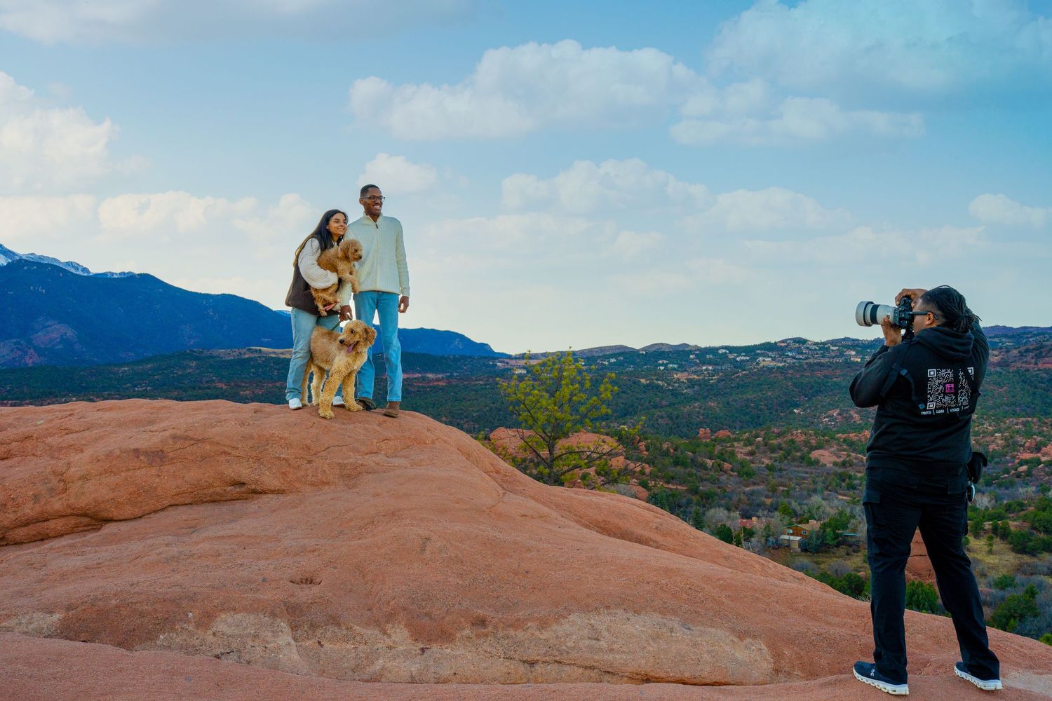 A photographer captures a couple and their dog posing on red rocks with mountain views in the background.