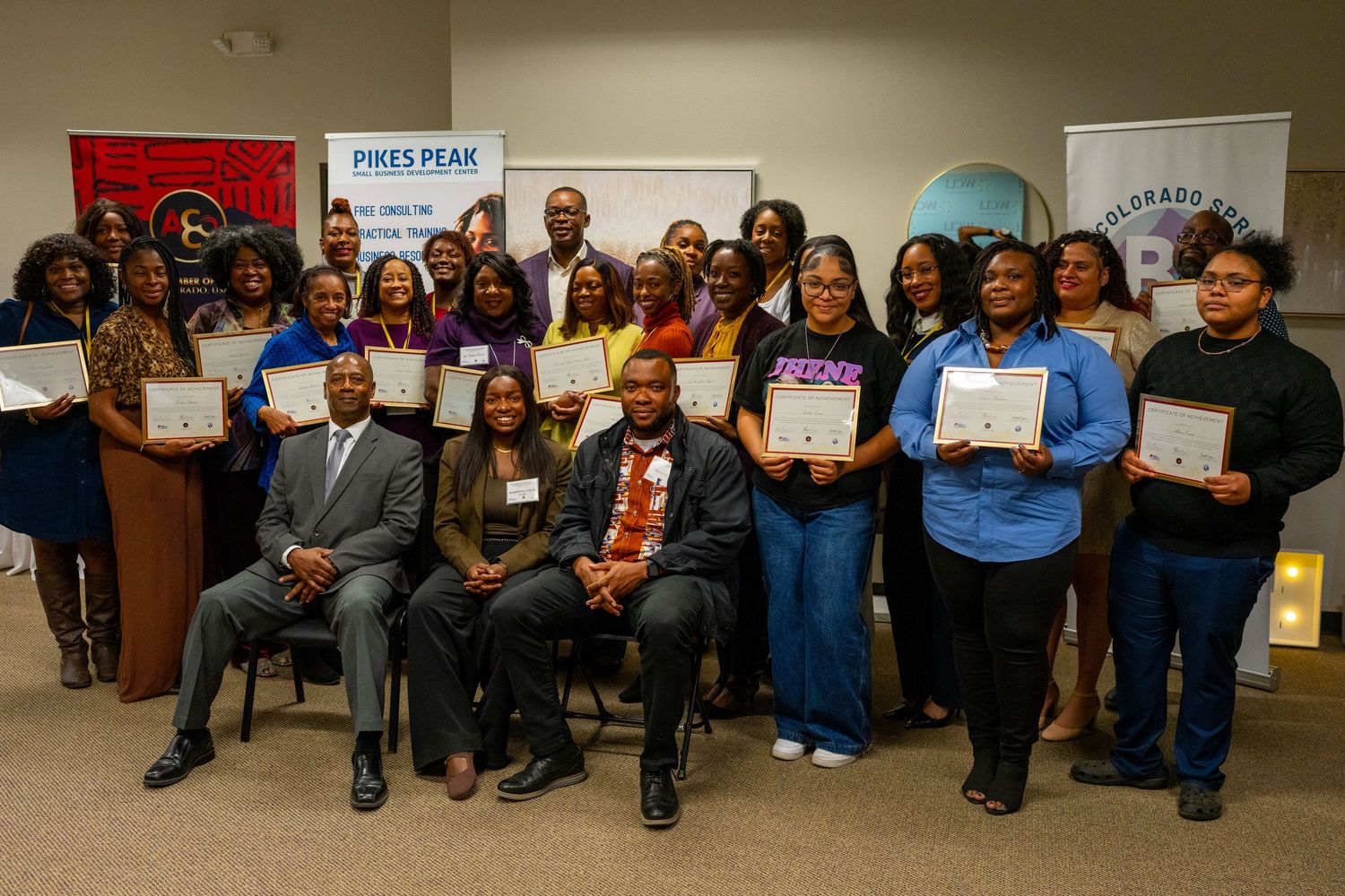 Group of people sitting together in a row at an indoor event wearing casual attire and holding certificates.