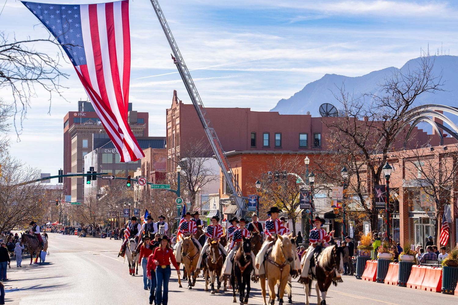 A parade procession with American flags and horses marches down a snowy small town street during a winter celebration.