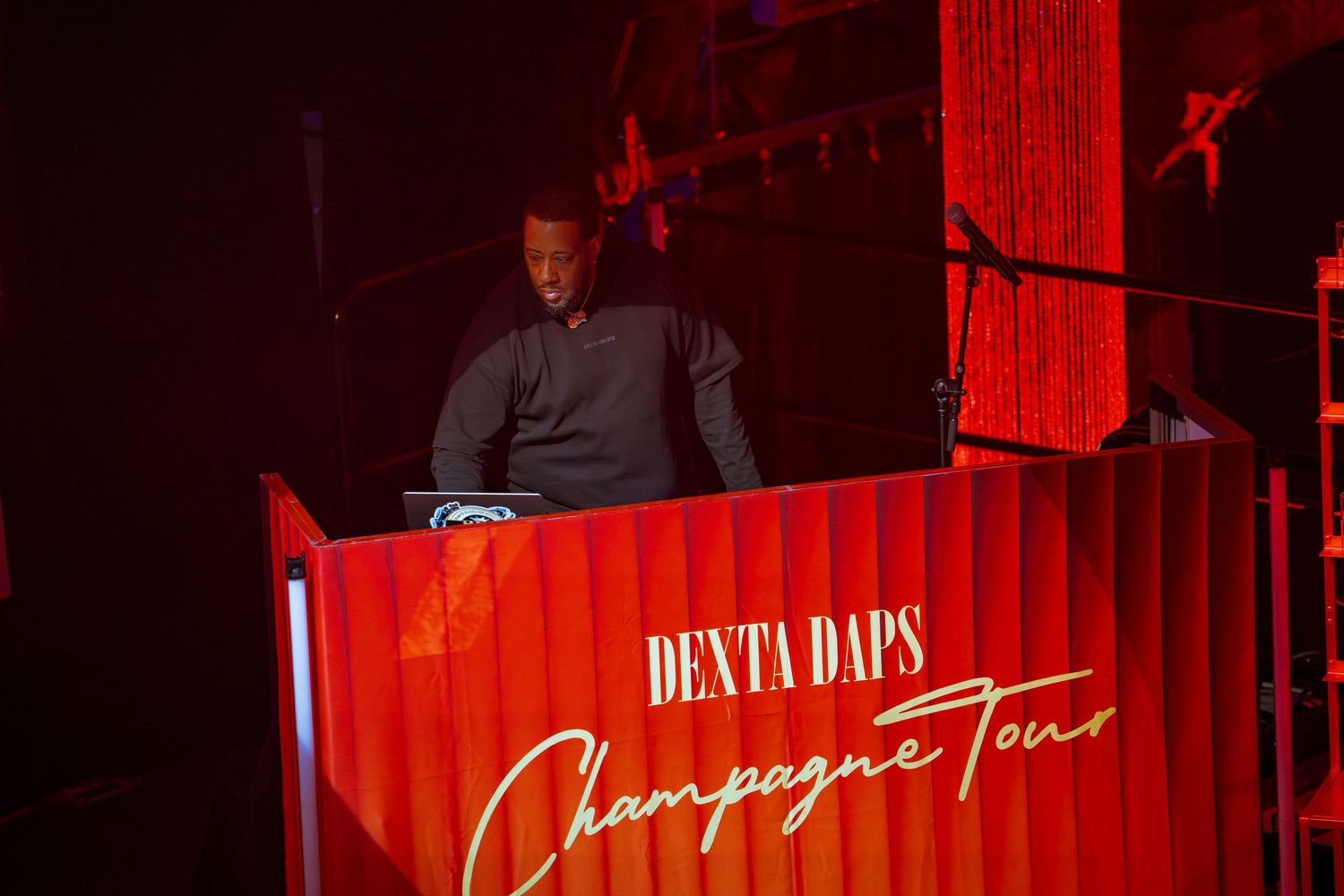 DJ performing at Dexta Daps Champagne Tour event on a red illuminated stage.
