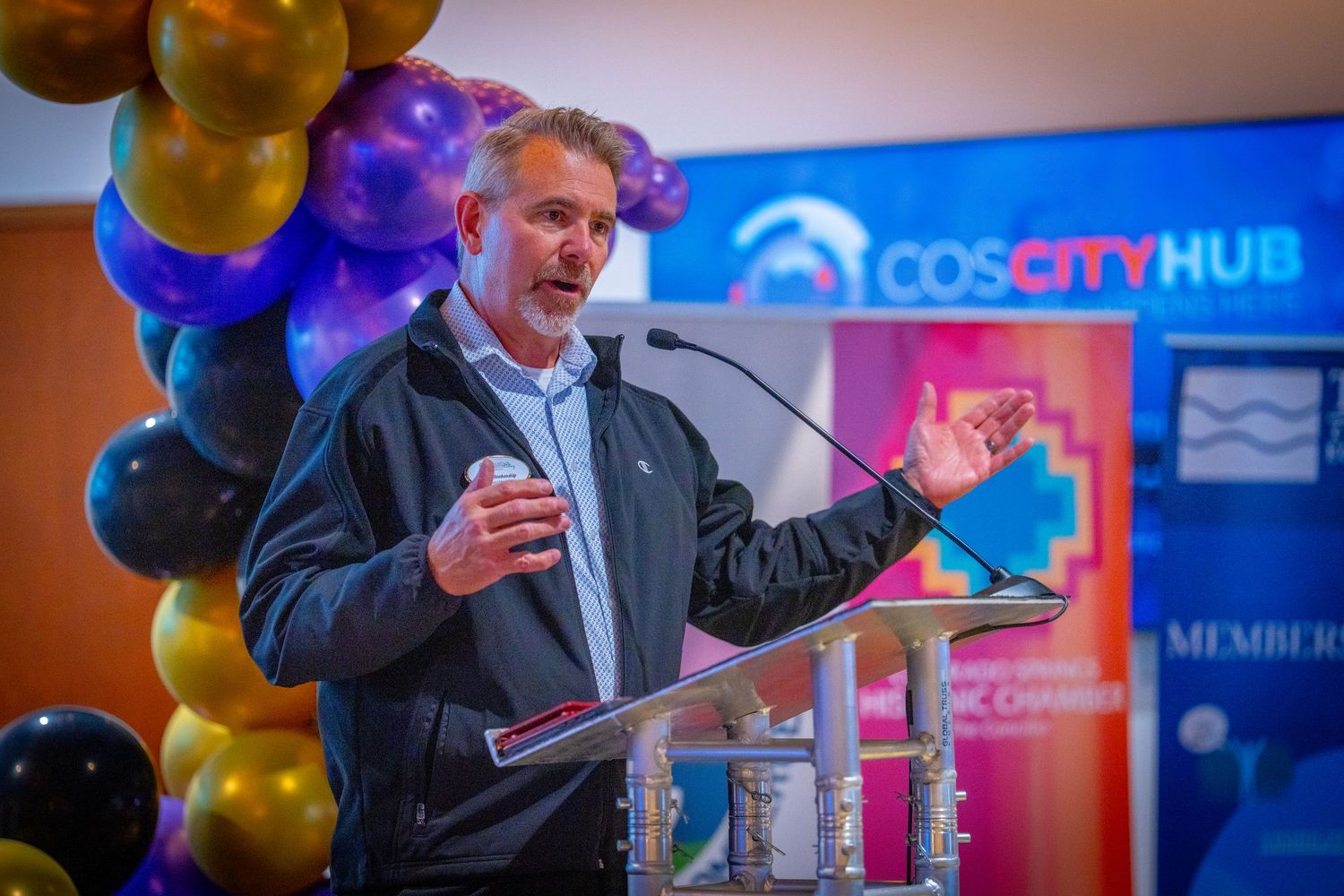 Man speaking at podium during COS City Hub event with colorful balloons in background.