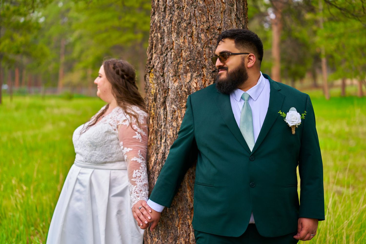 A couple poses by a tree trunk in wedding attire, with a green forest background.