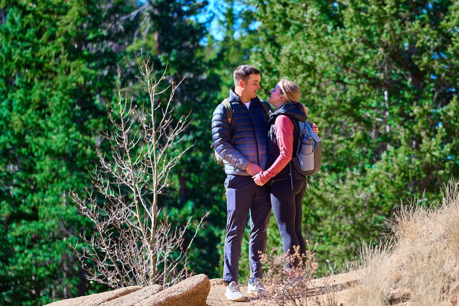 Couple embracing on trail against backdrop of evergreen forest.