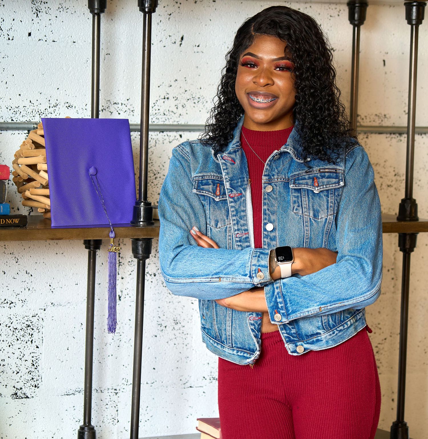 Person in denim jacket holds purple folder while posing against industrial wall background.