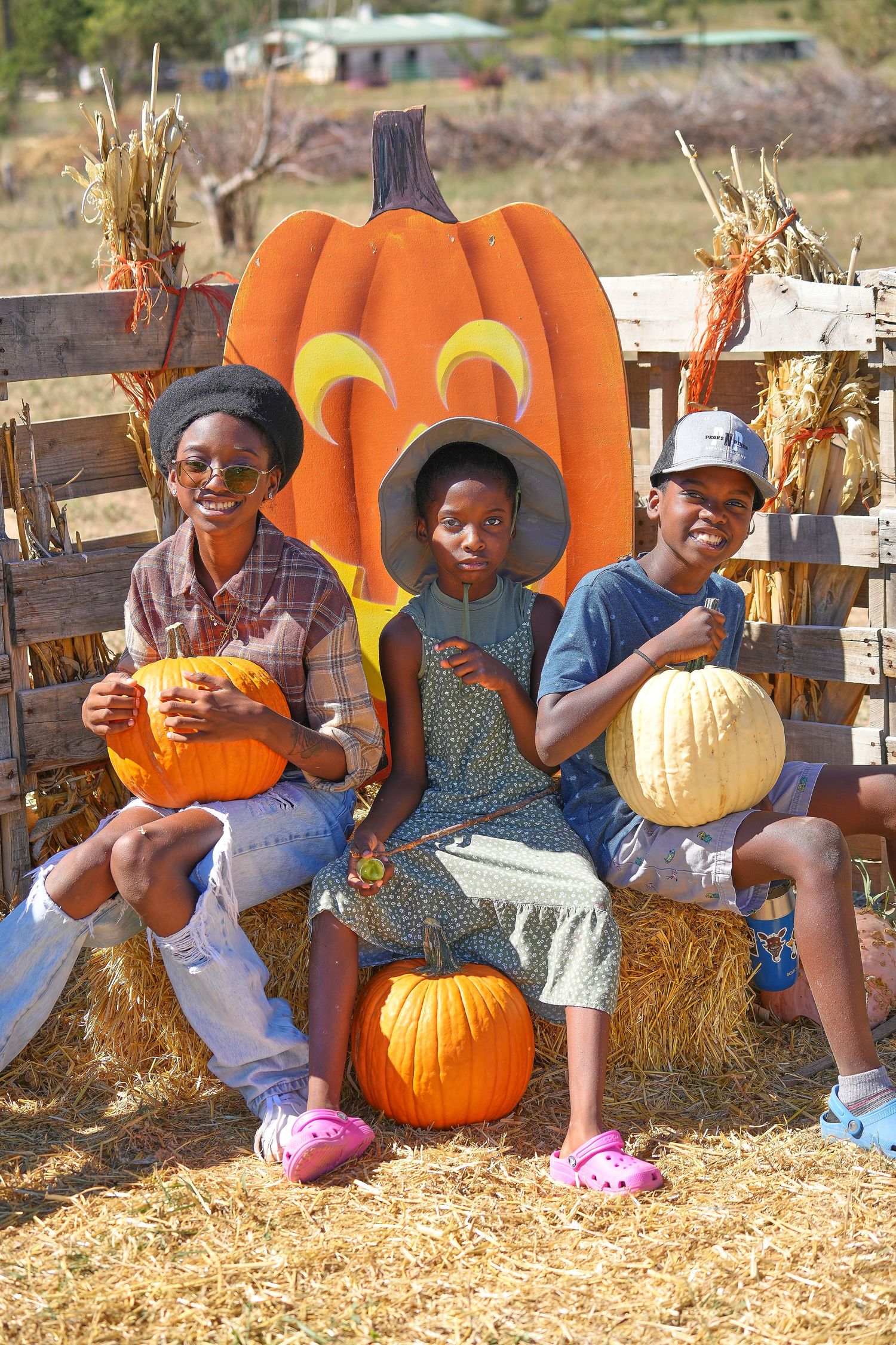 Three children pose with pumpkins on hay bales in front of large decorative orange pumpkin display.