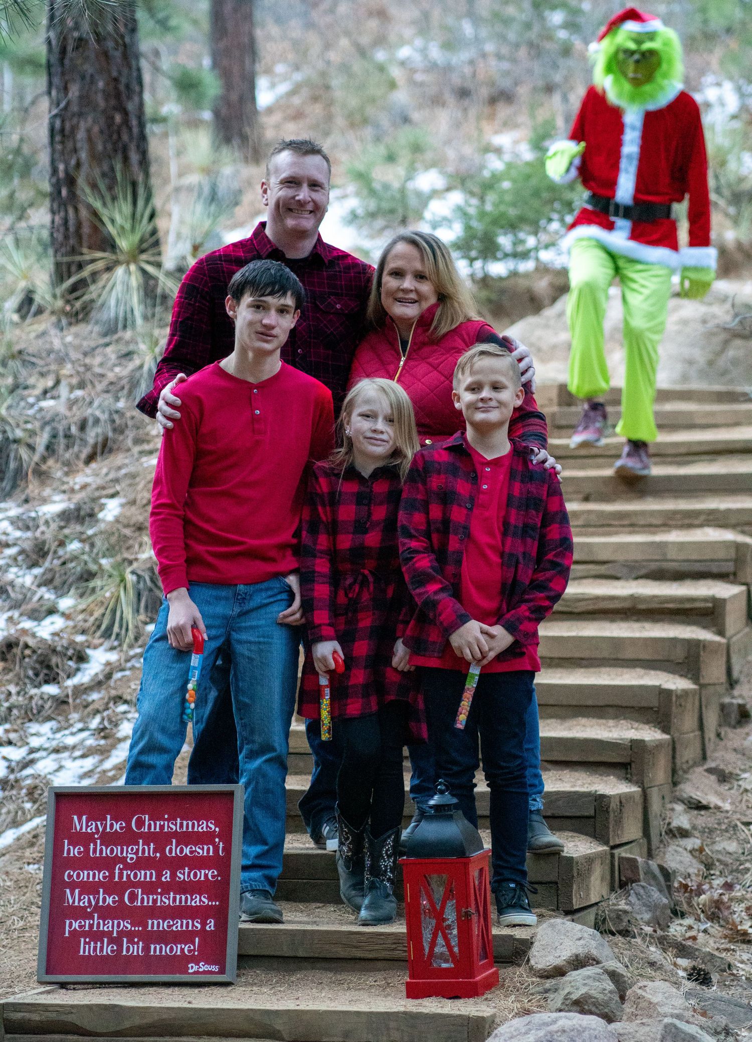 Family in red holiday outfits on stone steps with Grinch character lurking in background.