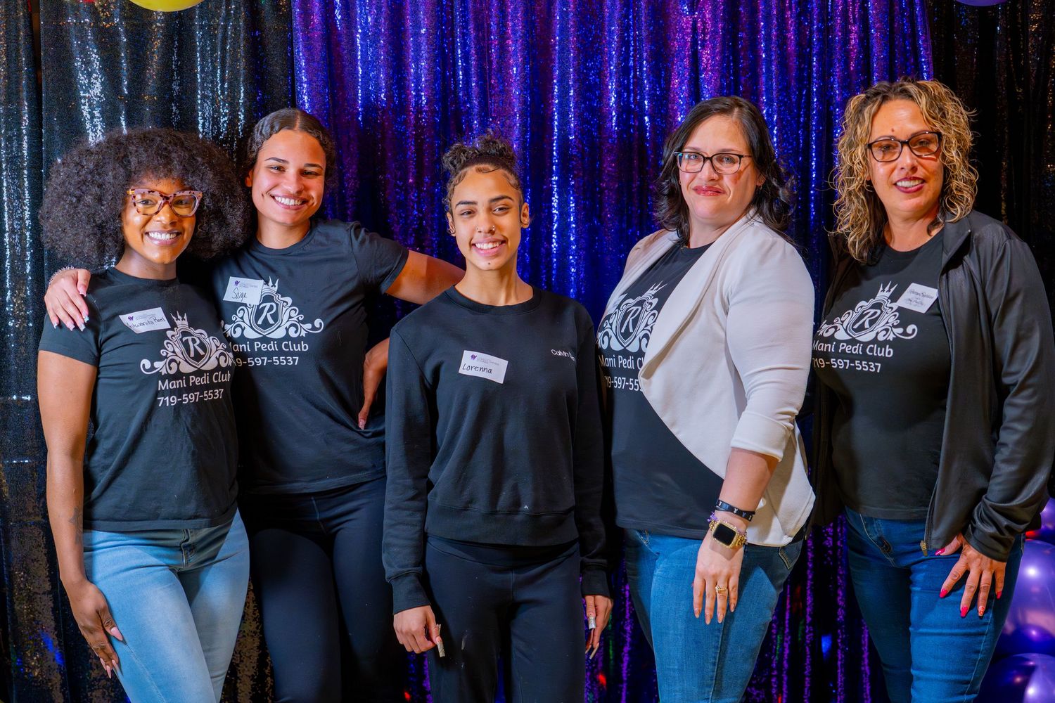 Five women smiling together in front of a sparkly backdrop, wearing matching black t-shirts at a group event.
