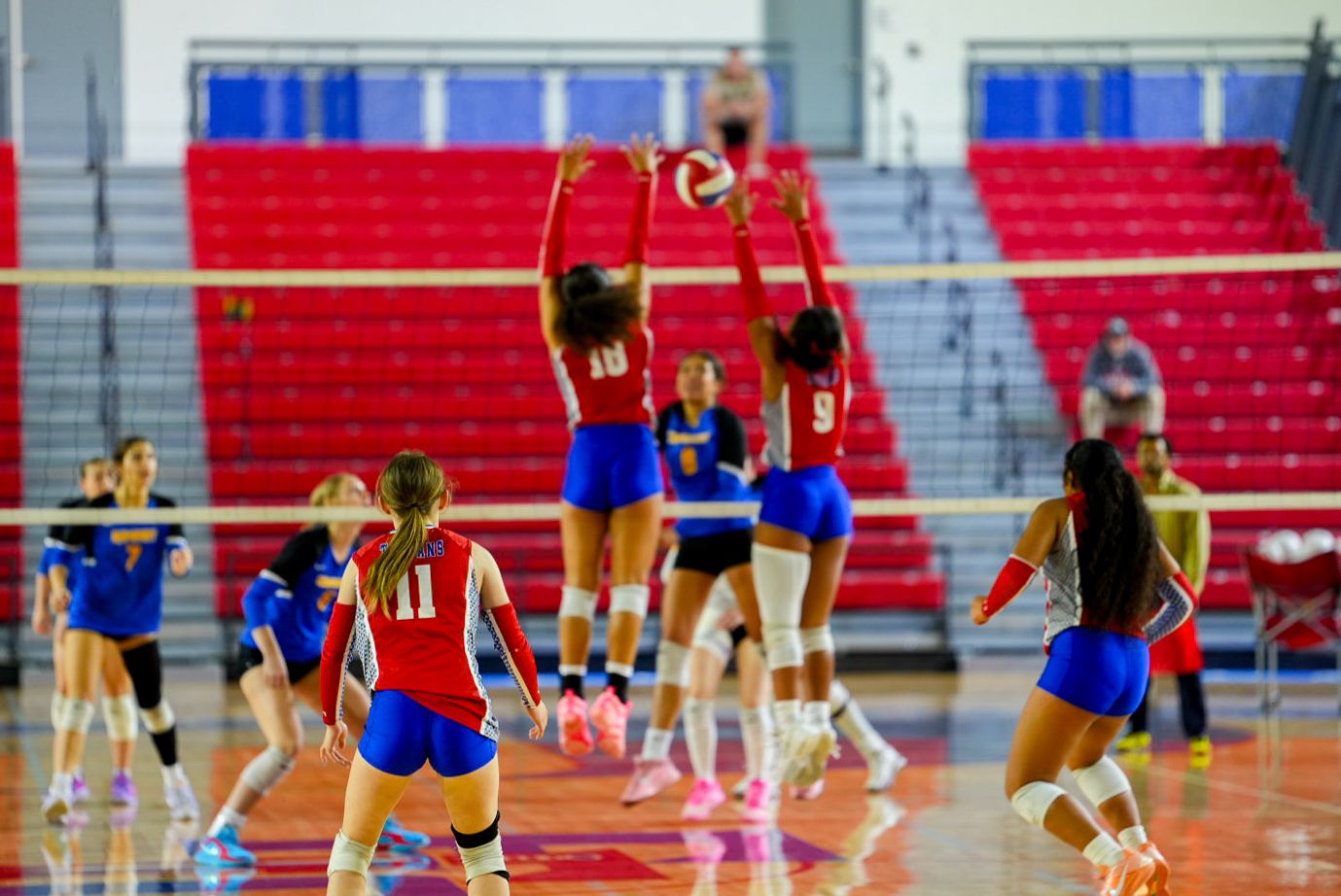Female volleyball players in red and blue uniforms compete during an indoor match with red bleachers in background.