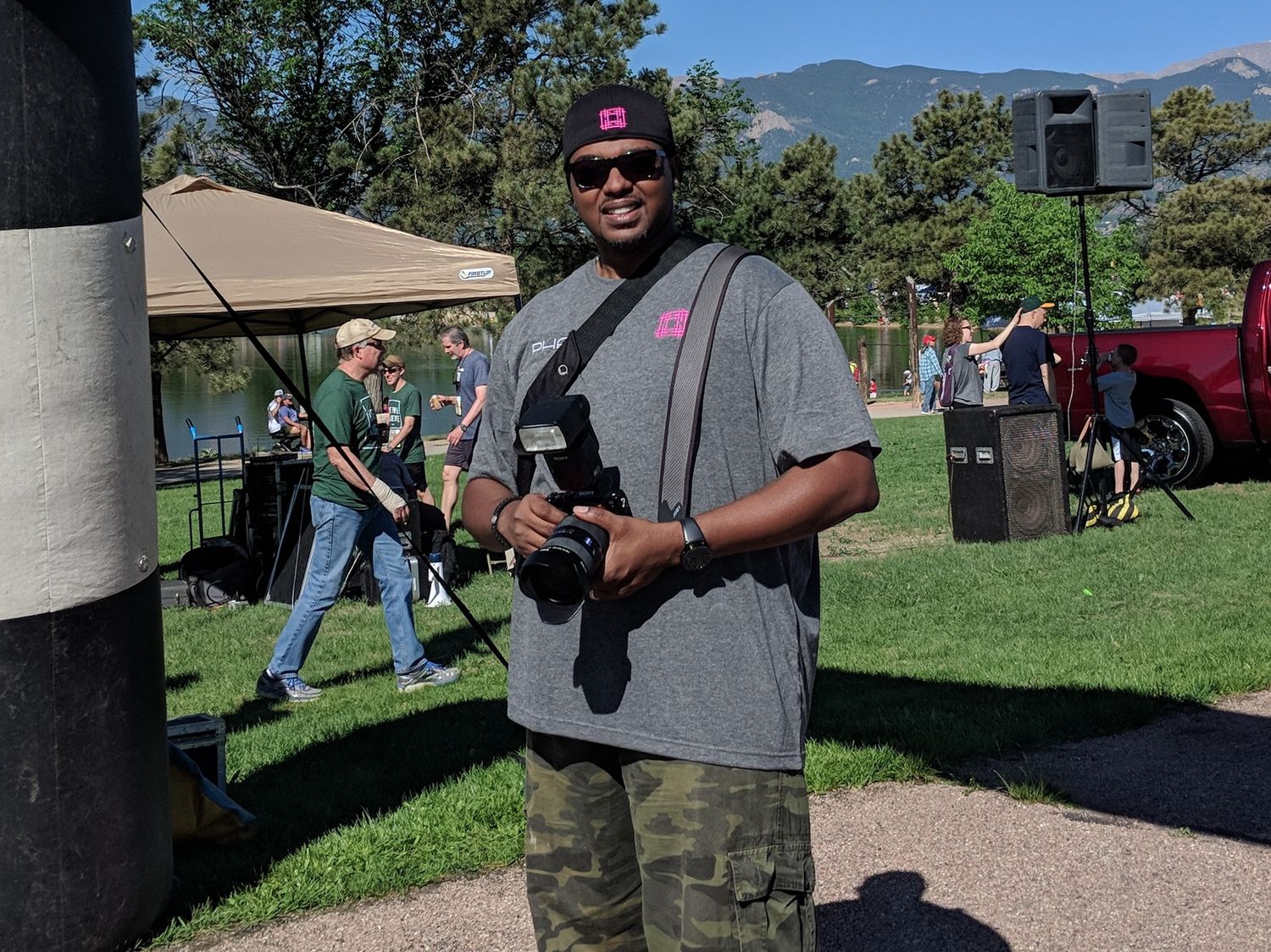 Photographer wearing camo pants and gray shirt stands outdoors with camera gear and mountains in background.