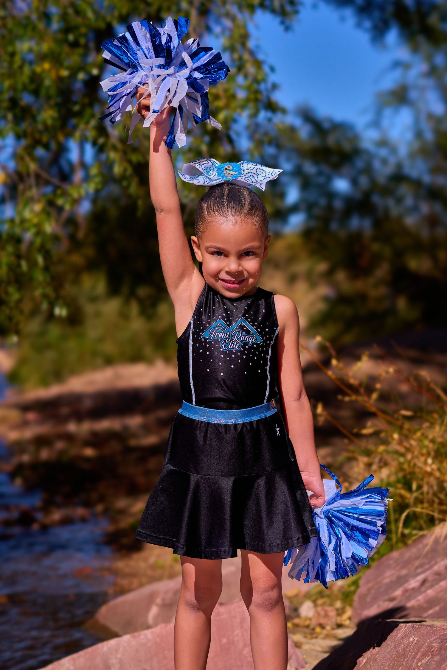Young cheerleader in a black and blue sparkly uniform with pom poms poses outdoors by a creek on a sunny day.