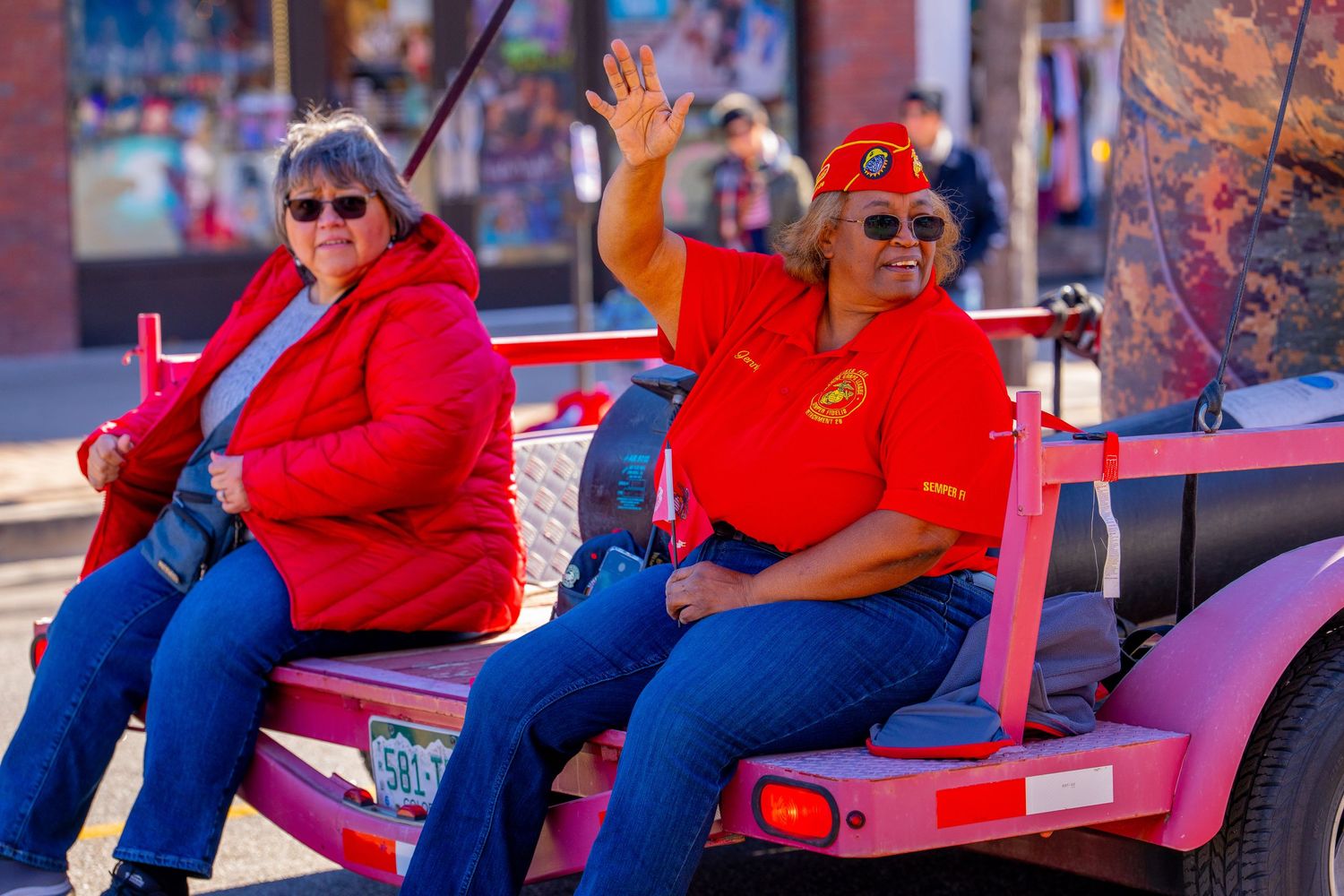 Two people in red shirts and blue pants wave while riding in a pink parade vehicle.