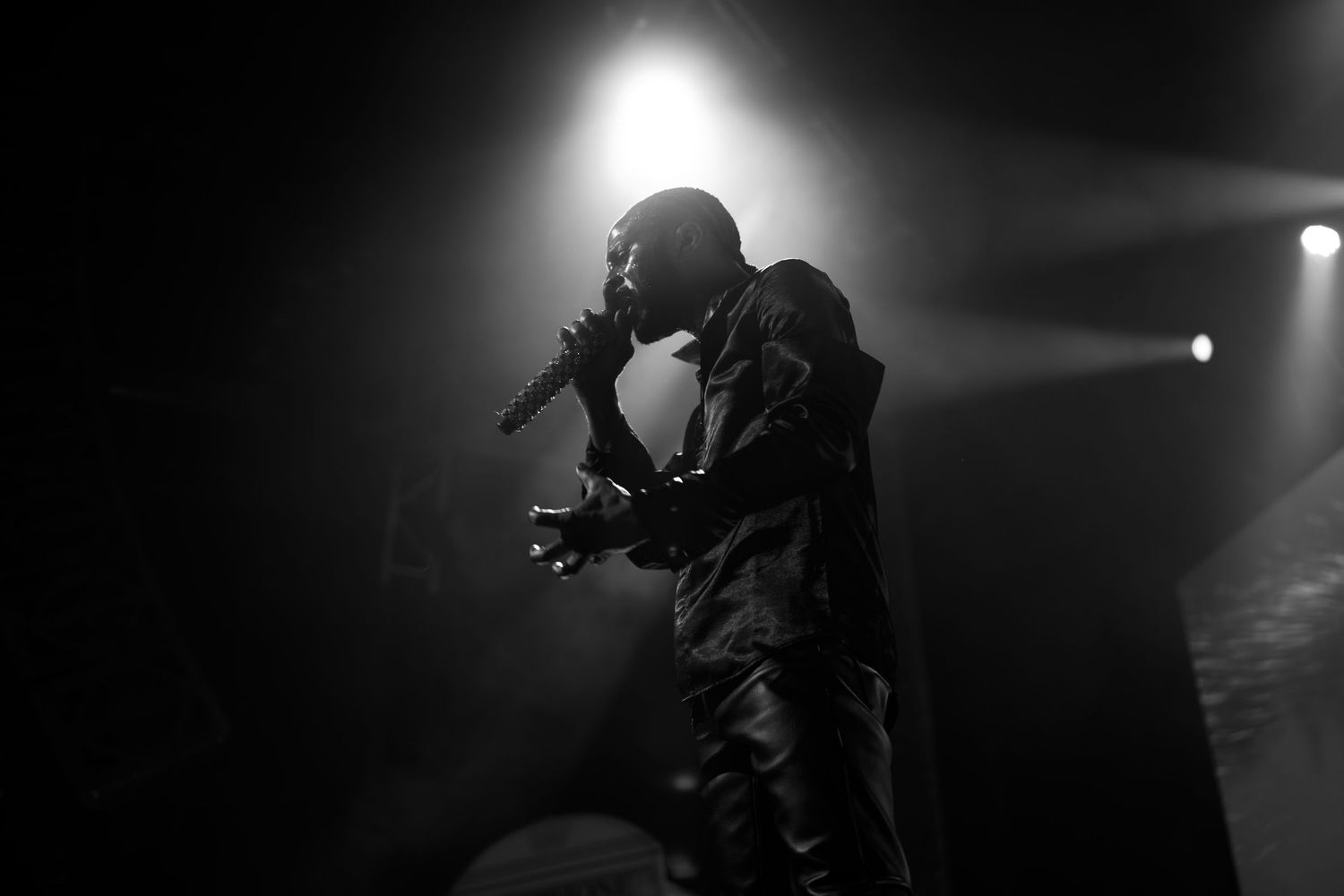 Black and white photo of a male performer singing into a microphone on a dark, fog-filled concert stage.