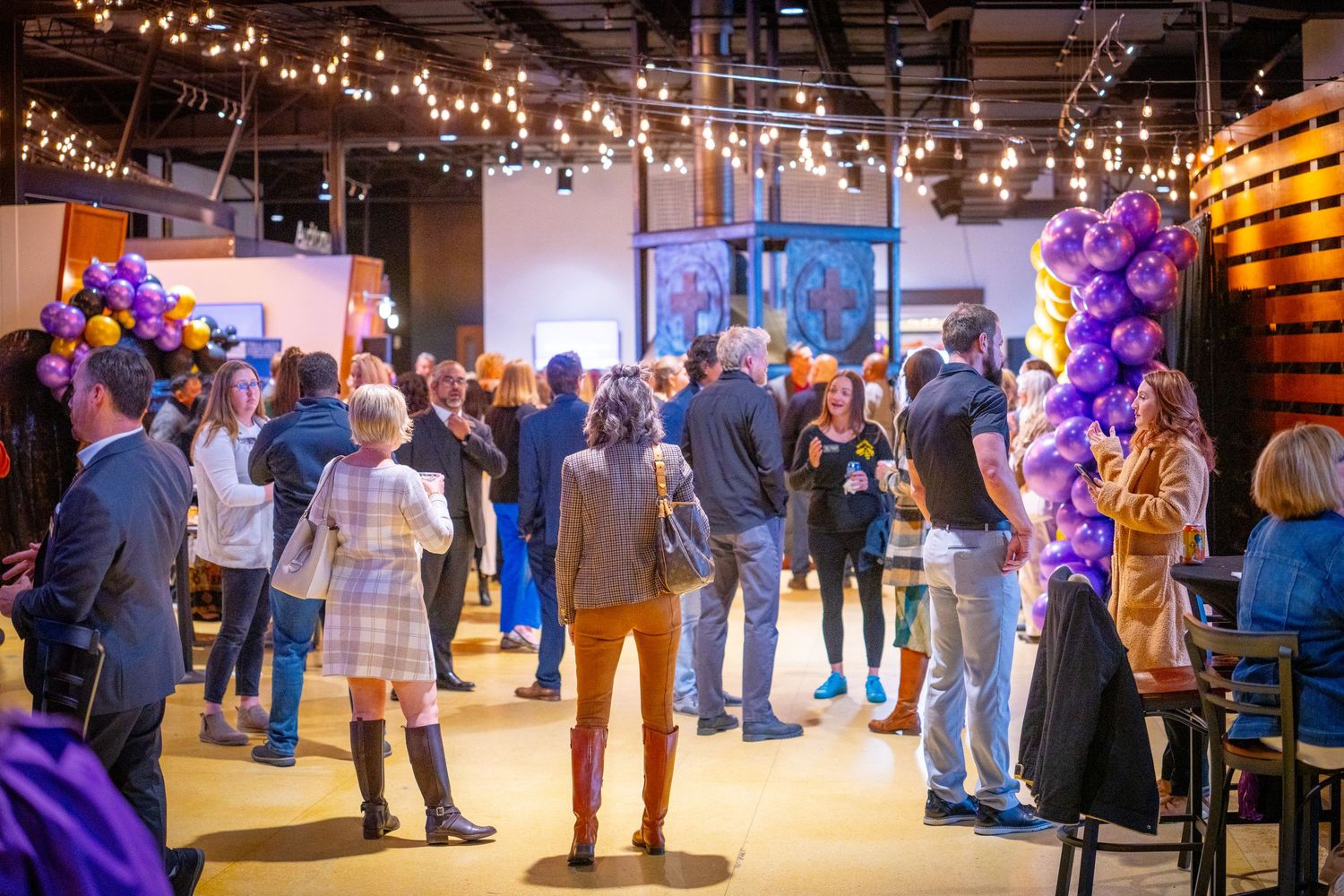 Guests mingle at a festive indoor event venue with string lights and purple balloon decorations.