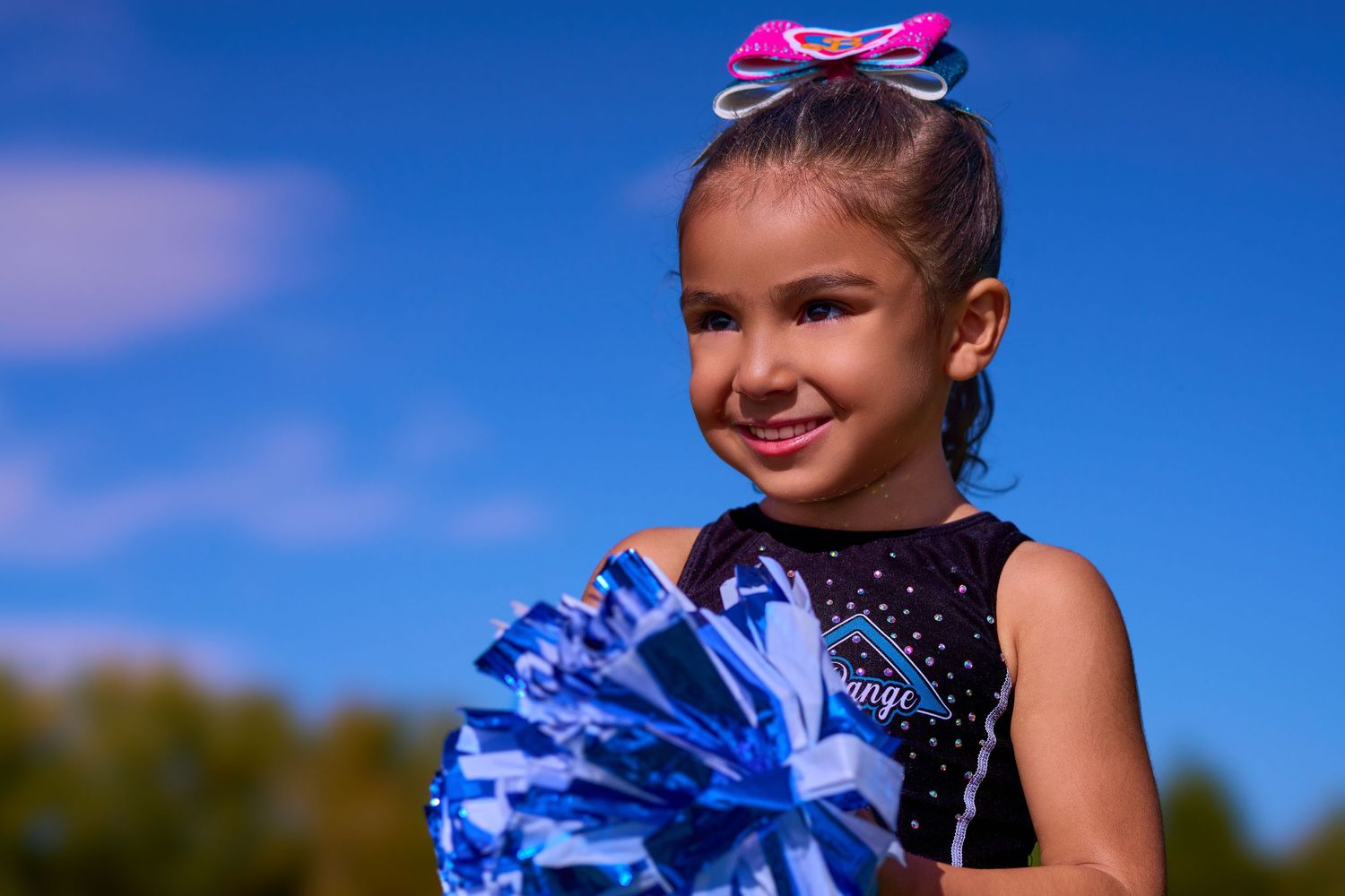 Cheerleader in sparkly blue and black outfit holds pom pom against bright blue sky background.