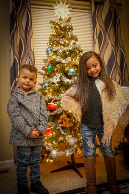 Two children posing in front of an illuminated Christmas tree with ornaments and decorations.