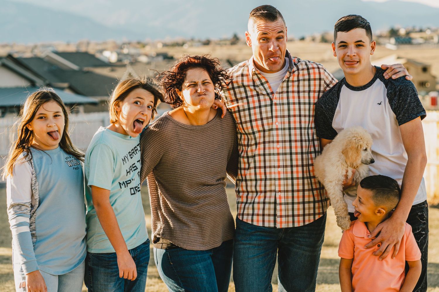A family of six posing together outdoors against a backdrop of rooftops and mountains at sunset.