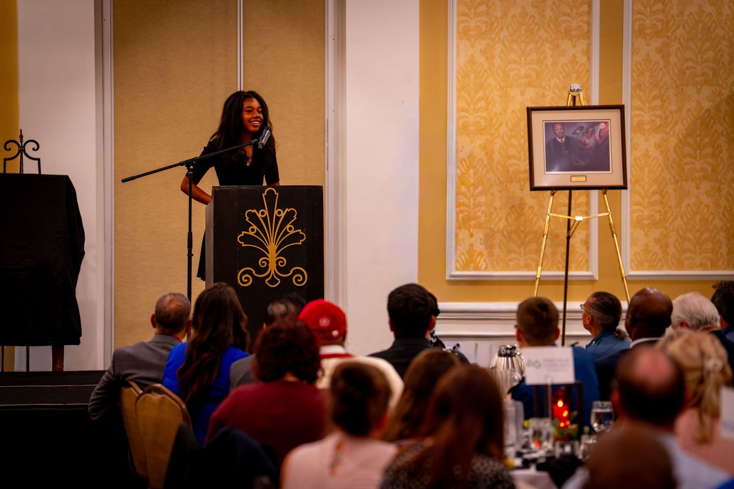Woman speaking at podium during formal banquet event in elegant ballroom with seated audience and artwork on easel.