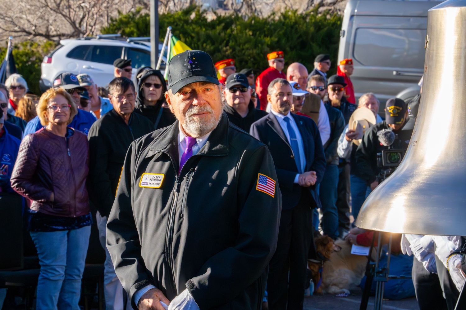 A crowd gathers at an outdoor event with a veteran wearing a black vest with American flag patch in the foreground.