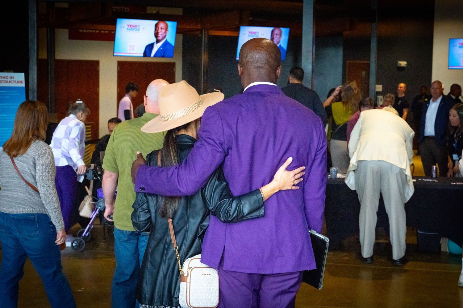 Man in purple suit with arm around woman in wide-brim hat at a political event with TV screens in background.