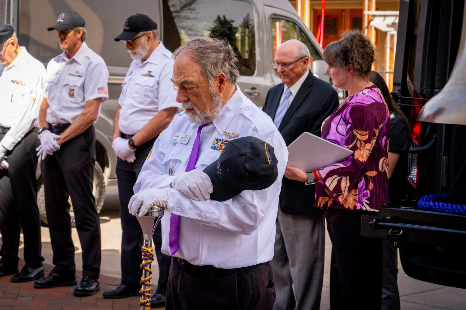 Veterans in white uniforms bow heads during a solemn outdoor memorial ceremony as a woman reads from papers nearby.