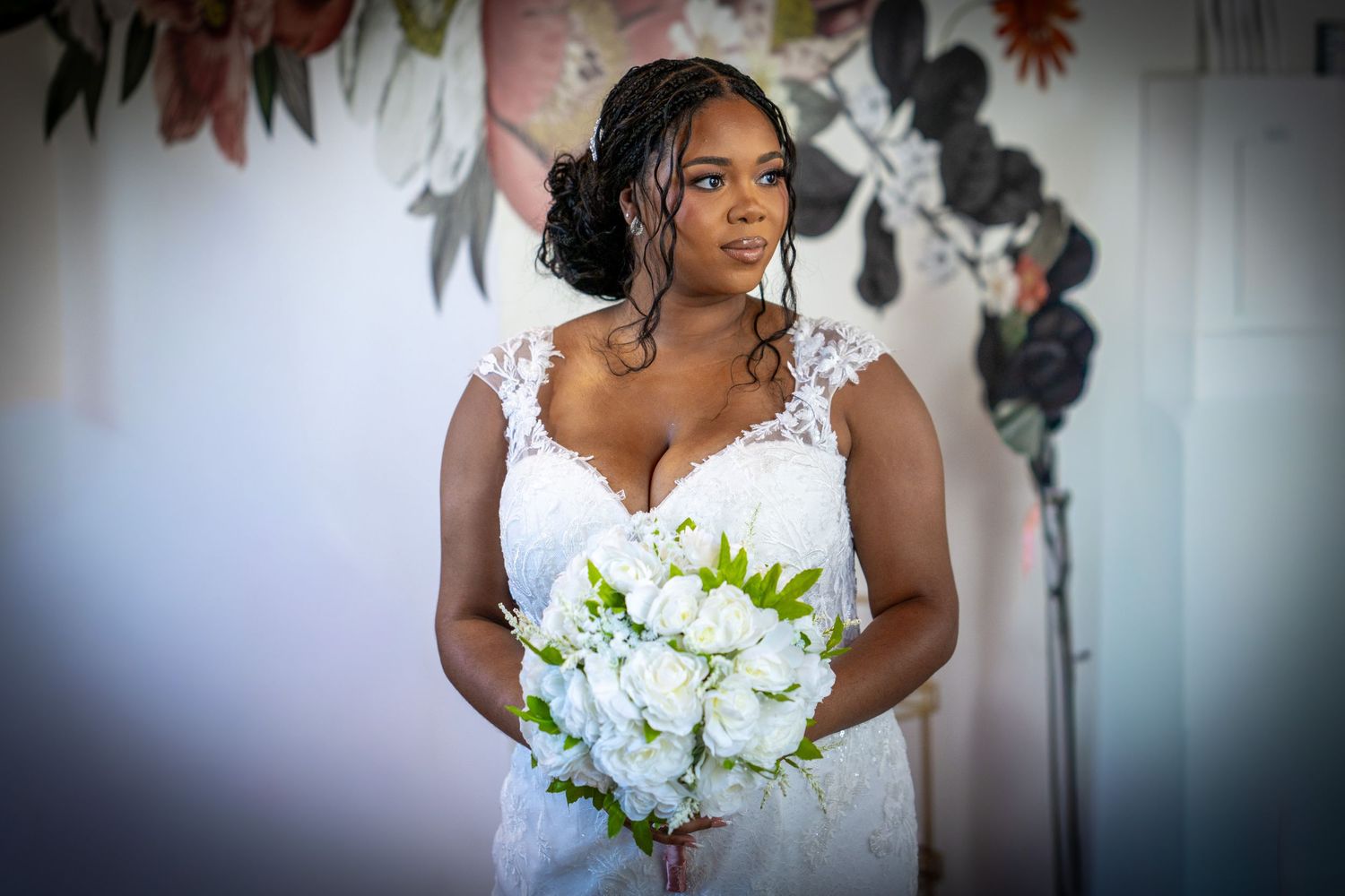 Bride in white lace gown holding white rose bouquet, standing before floral backdrop on wedding day.