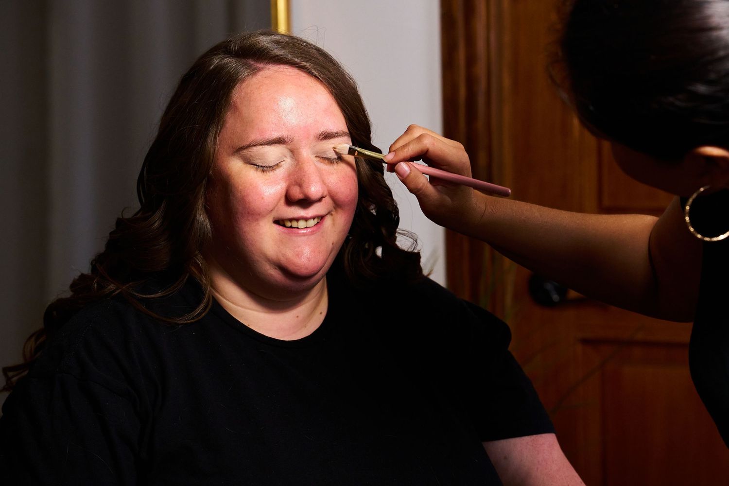 Someone getting their makeup professionally applied while smiling with eyes closed.