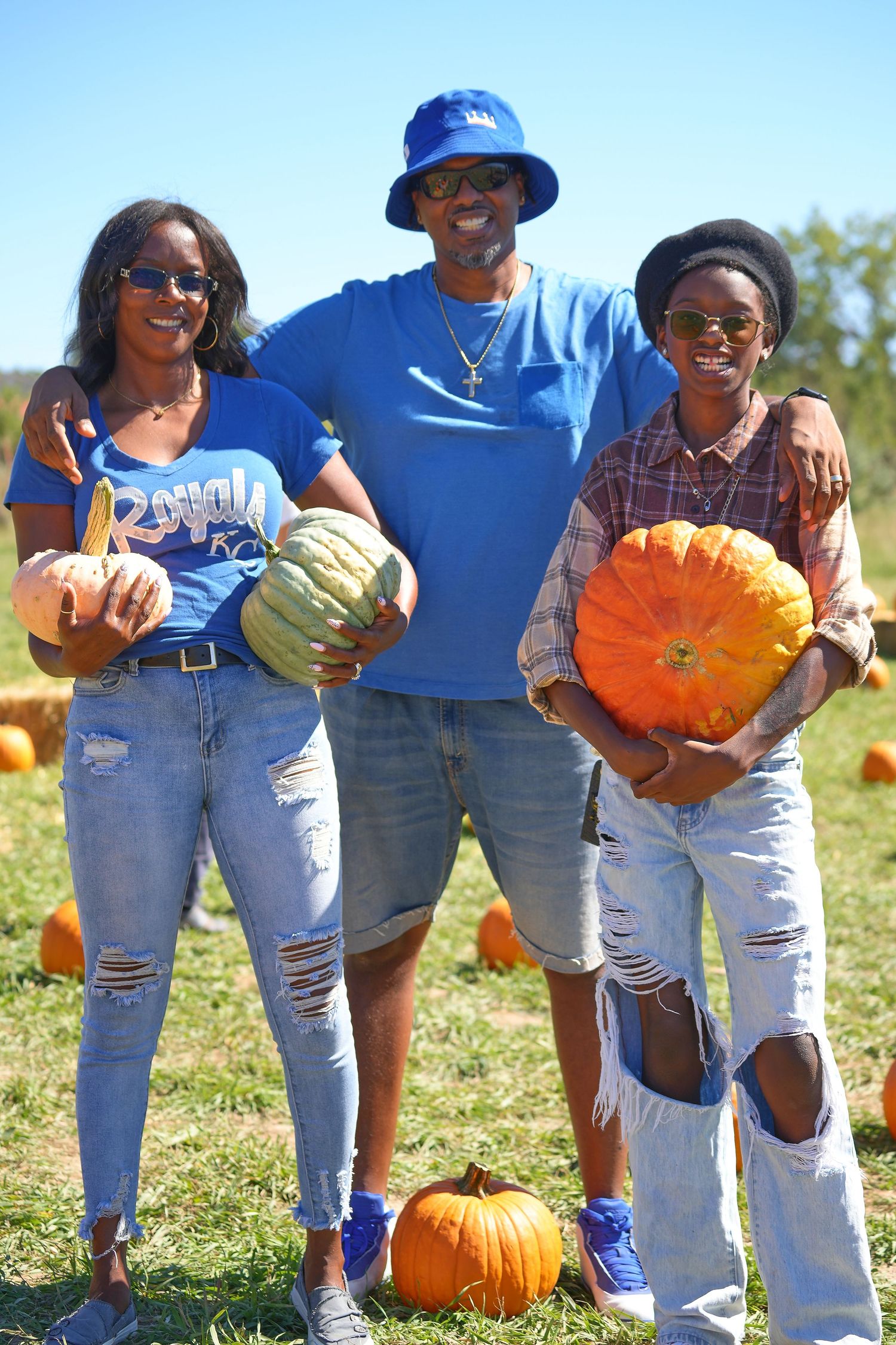 Three people in casual wear hold pumpkins while posing at outdoor pumpkin patch.