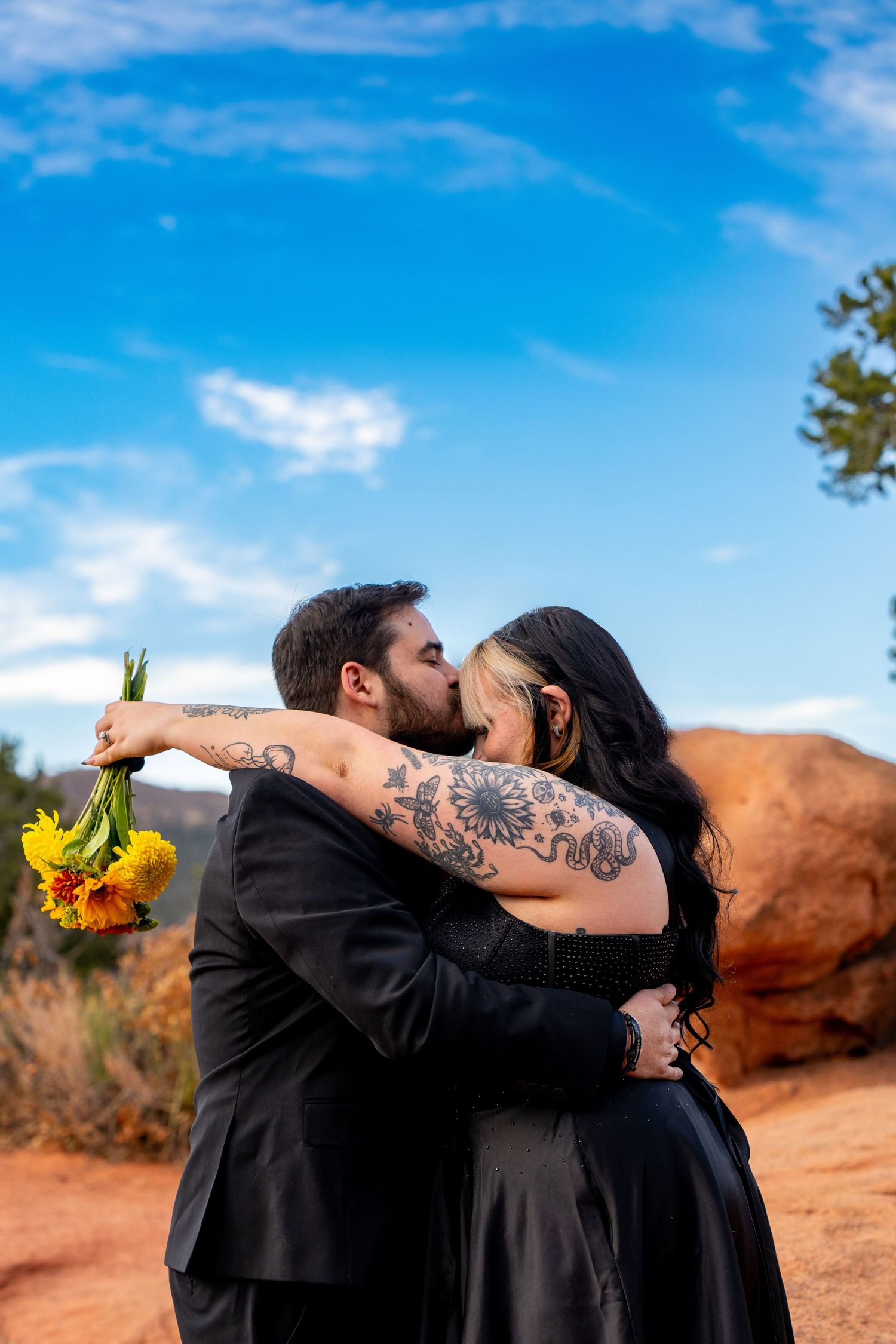 A couple in black attire share a romantic kiss in front of red rocks and blue sky while holding sunflowers.