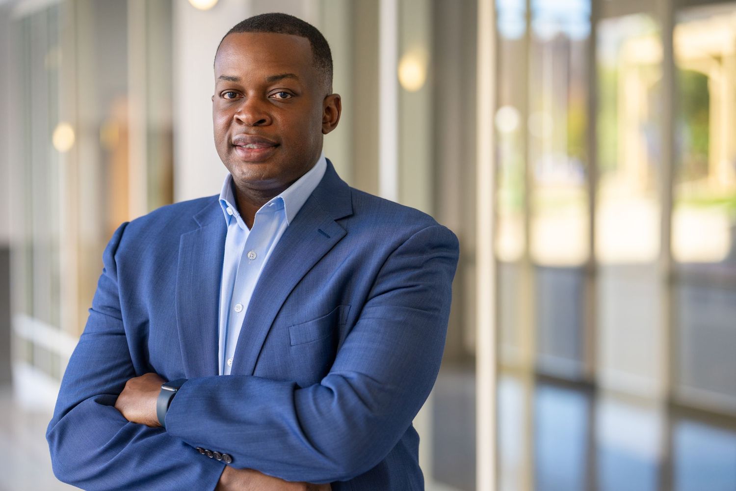 Professional wearing a navy blue suit and light blue tie poses in a modern office hallway with glass walls.