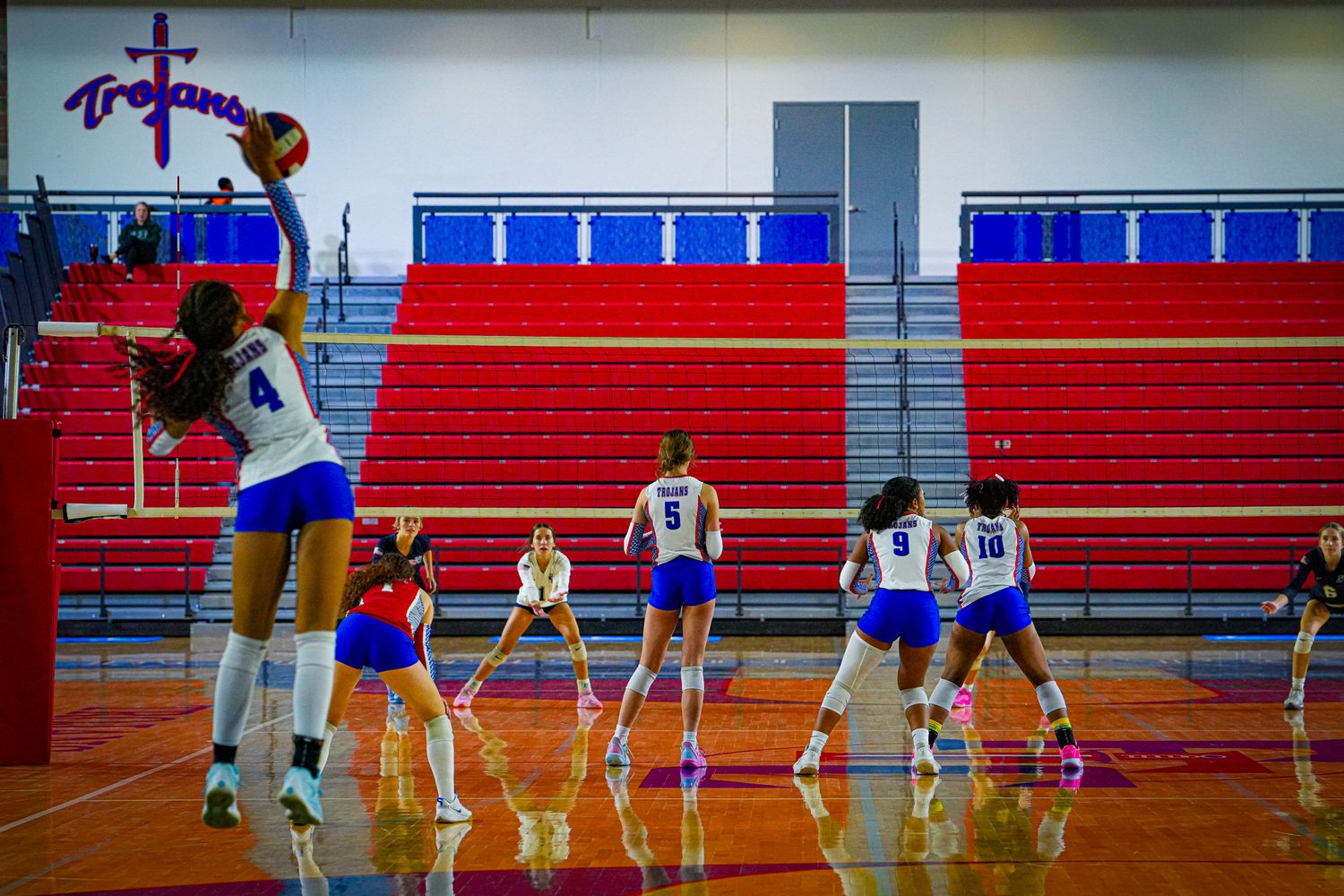 Female volleyball players in blue uniforms compete in a gym with red bleachers and a Trojans banner during a match.