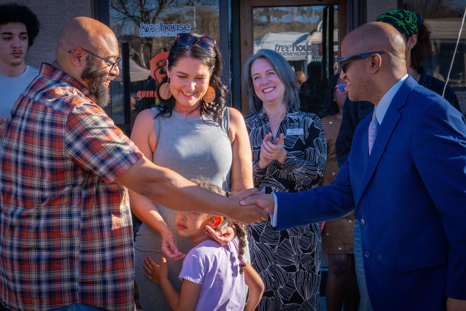 People shaking hands and smiling at an outdoor community event near a storefront on a sunny day.