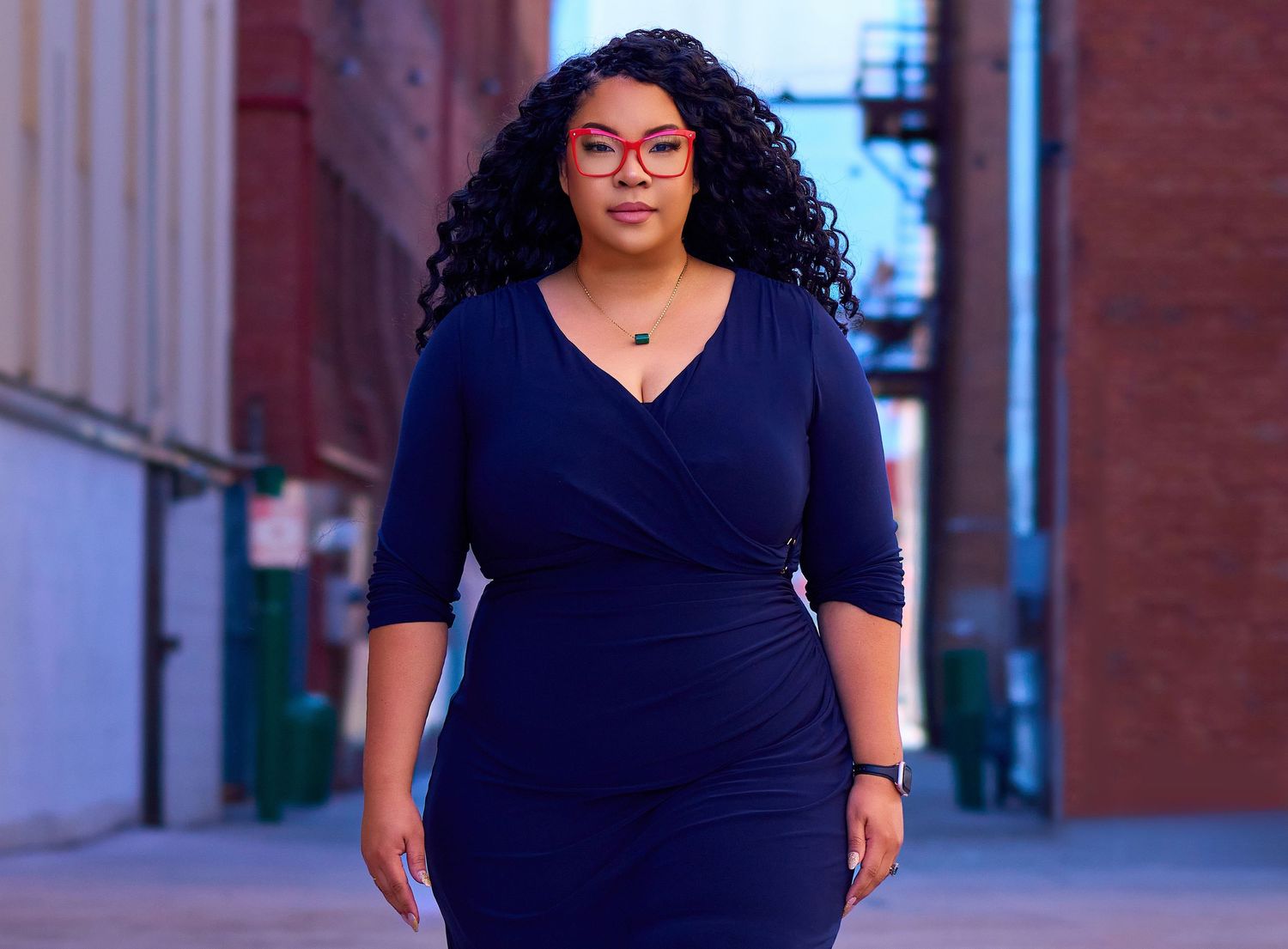 Professional wearing a navy blue wrap dress with glasses stands confidently in an urban setting.