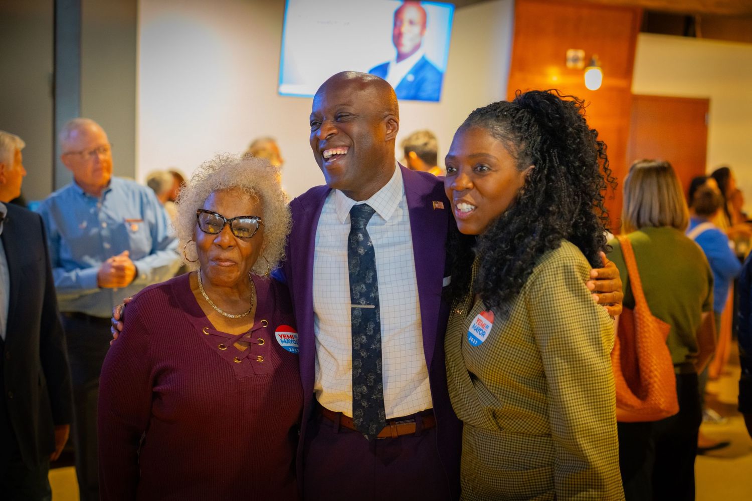 Three people smiling and posing together at a political campaign event, wearing name tags in a bright indoor venue.