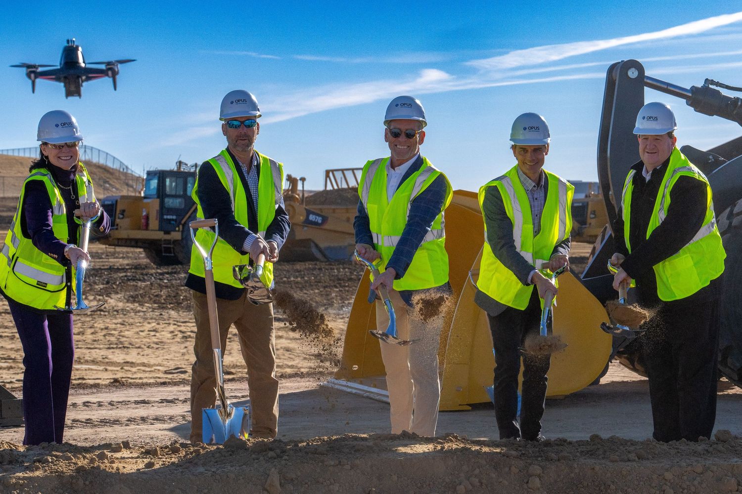 Five professionals in safety vests and hard hats participate in a groundbreaking ceremony with golden shovels at a construction site.